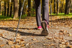 A person with wide feet is walking in a park using trekking poles. The scene includes fallen leaves on a gravel path, surrounded by trees under sunlight. The focus is on the person's legs and shoes as they stride forward, highlighting practical footwear tips for those with similar concerns.